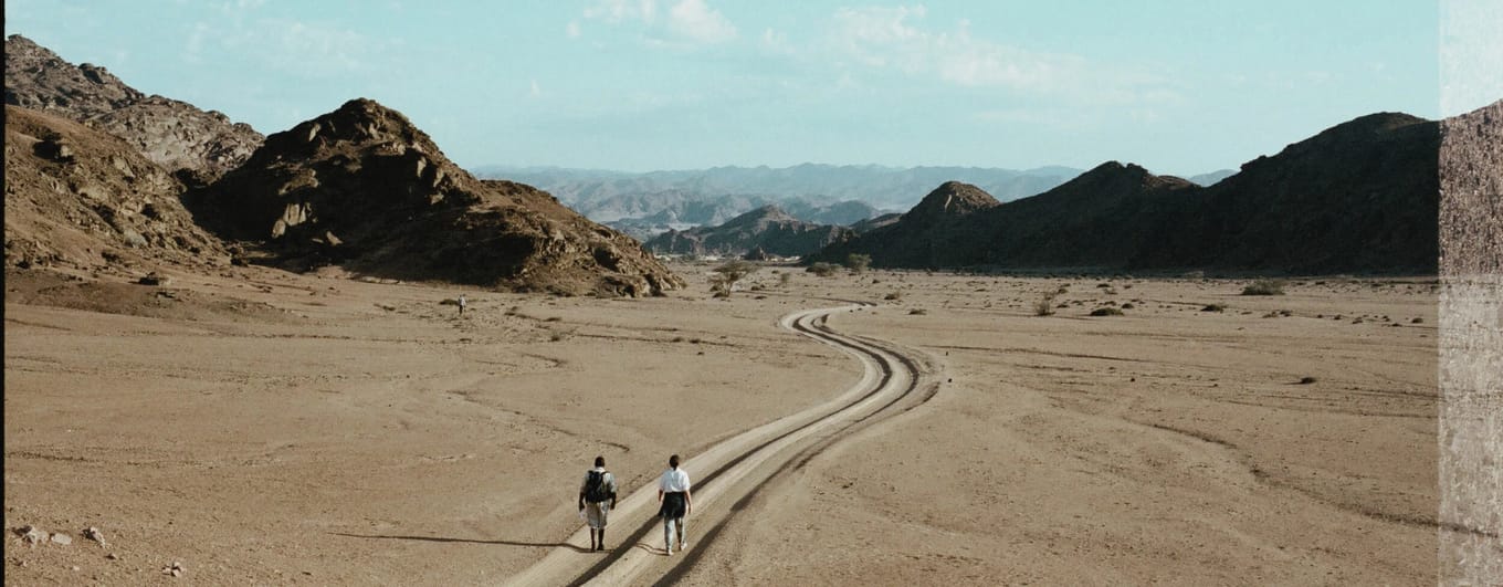 Man and woman walking along a dirt road in mountainous desert landscape